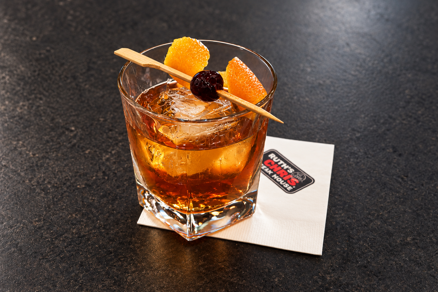 Ruth's Chris Madagascar Vanilla Old Fashioned over a large ice cube, garnished with orange peel and a cherry, on a Ruth's Chris cocktail napkin